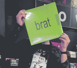 A heavily dithered two-quarters pose of a person holding a copy of the brat album on vinyl in front of their face in a record store. The person wears a checkered flannel over a band tee.